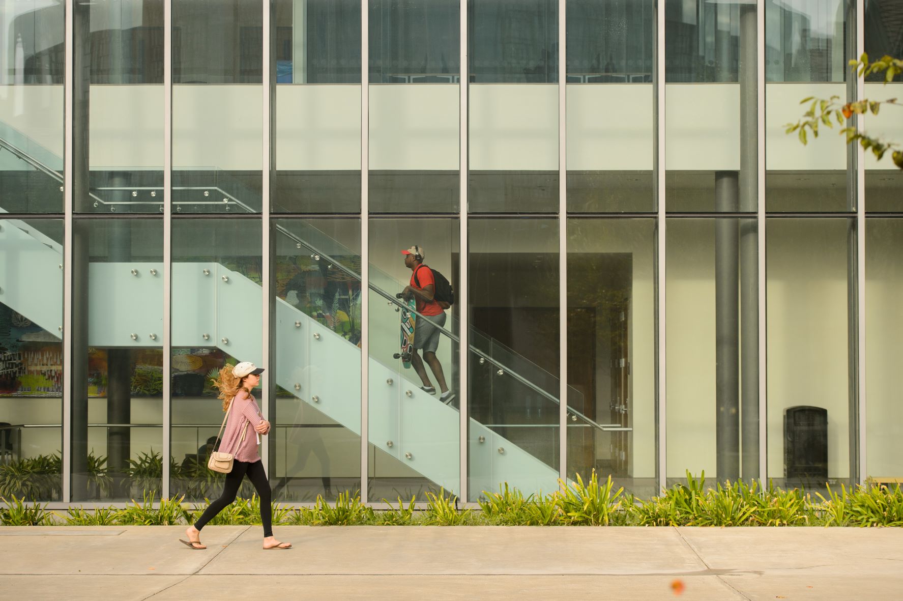 A glass wall separates two casually dressed students. One climbs stairs while the other walks past outside.